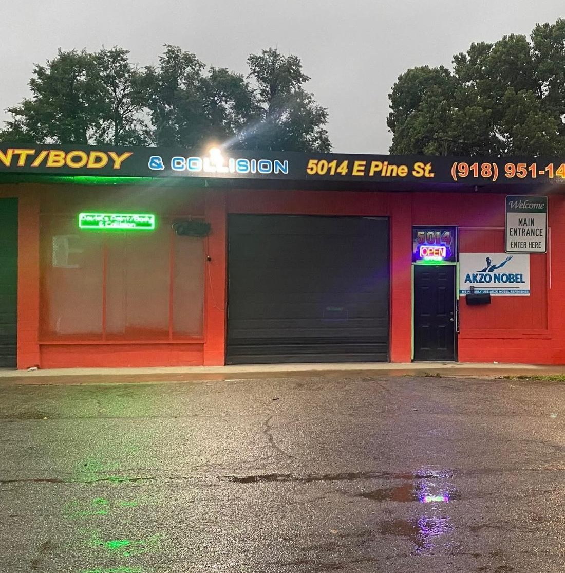 Daviel's Paint, Body & Collision at 5014 E Pine St in Tulsa. Neon signs lit at dusk, trees and overcast sky above, rain-slick parking lot in front